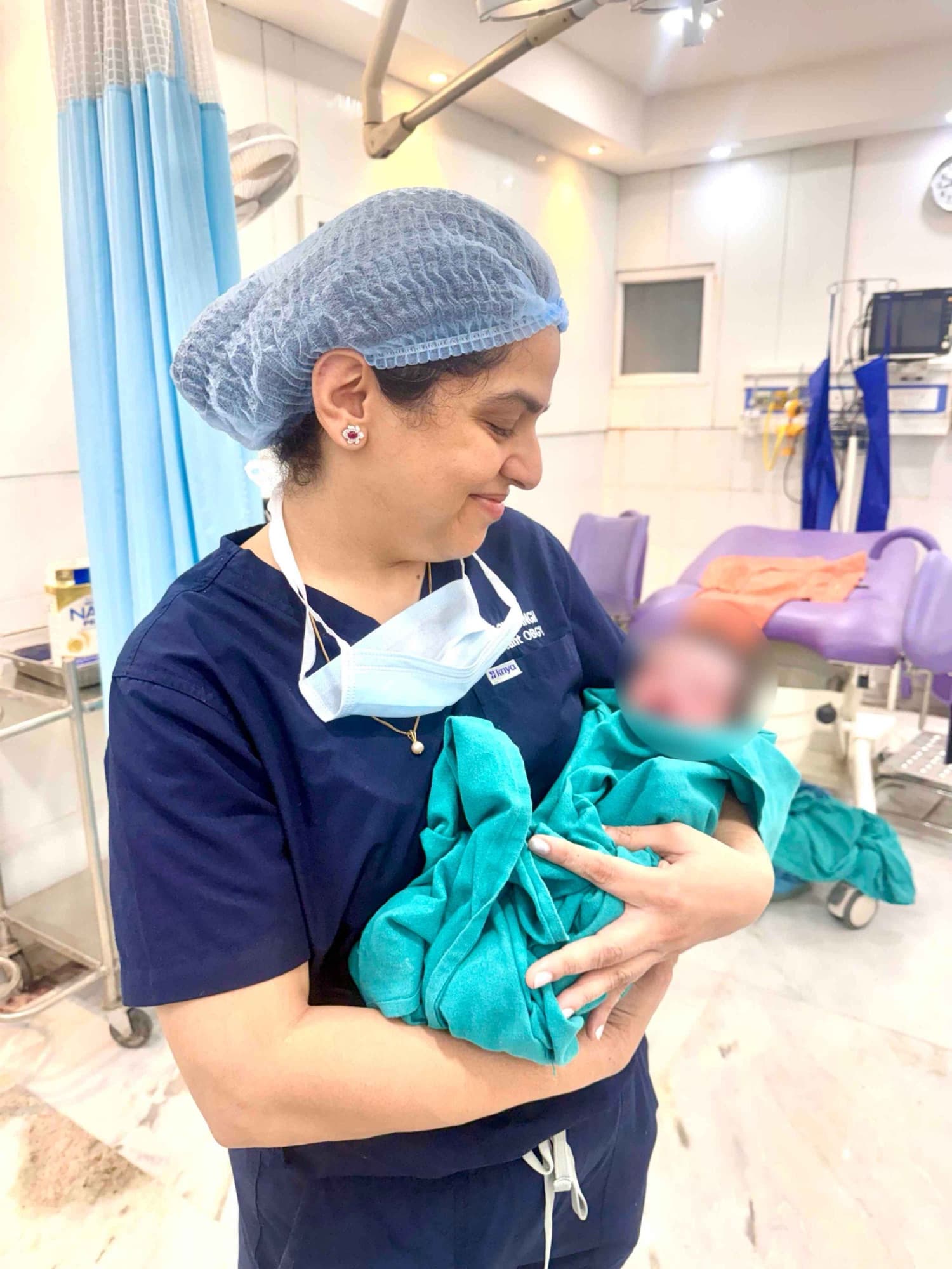 Dr. Shachi Singh smiling down at a newborn wrapped in a teal cloth in the delivery room at Prakash Hospital Noida