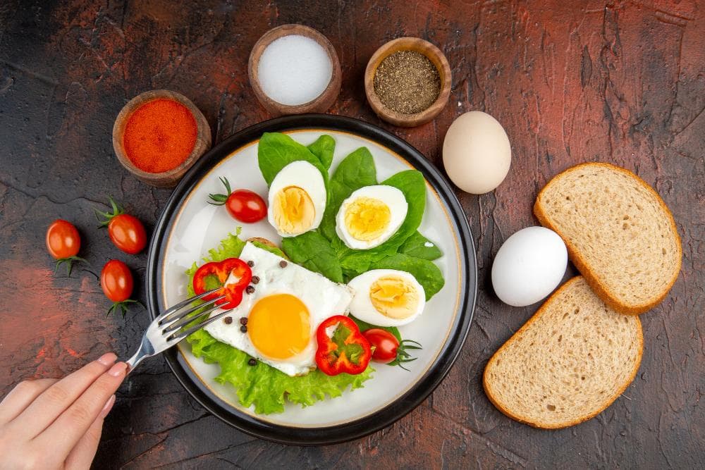 On a wooden table a healthy meal is shown, including eggs, tomatoes, bread, leafy greens, salt, pepper & chilli powder with focus on a woman's hand going to eat it with a fork in hand.