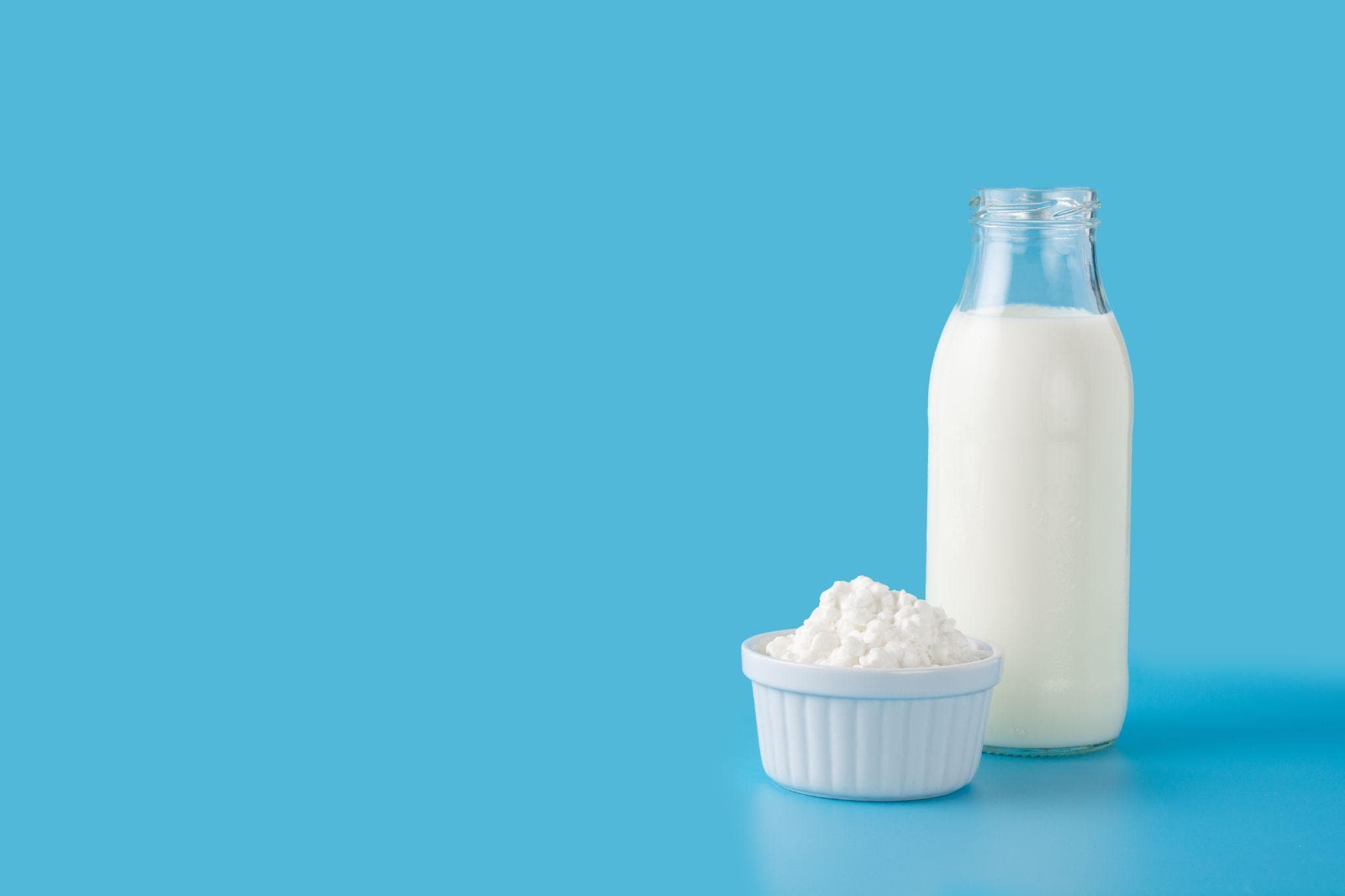 A glass bottle of milk and a bowl of cottage cheese are placed against a blue background.
