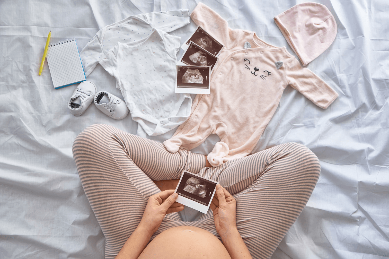 Pregnant woman looking at her fetus’s sonogram image with baby clothes and shoes arranged on the bed along with a notepad.