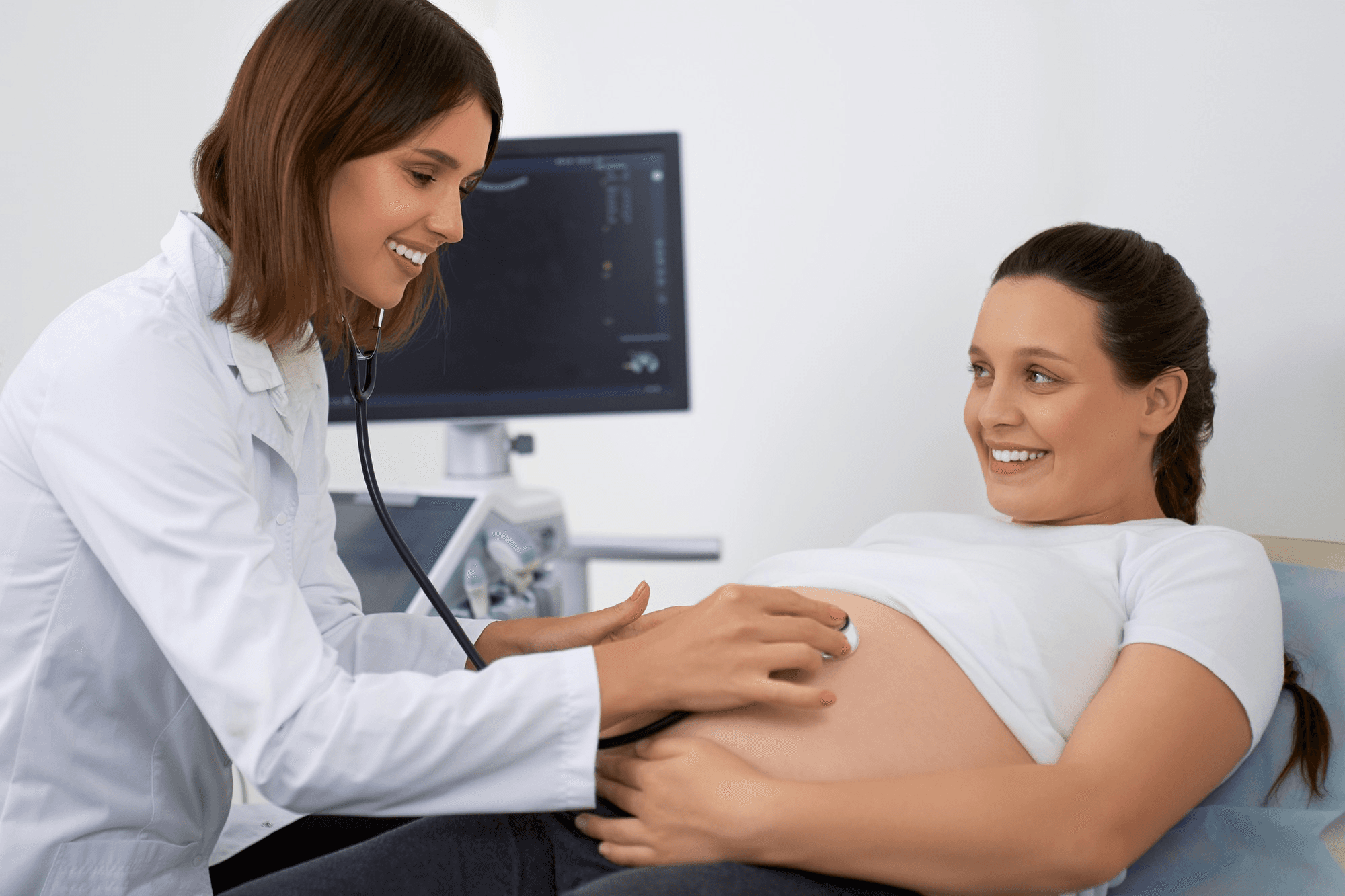Pregnant woman having an ultrasound performed on her belly by a health professional, while smiling at the ultrasound screen.