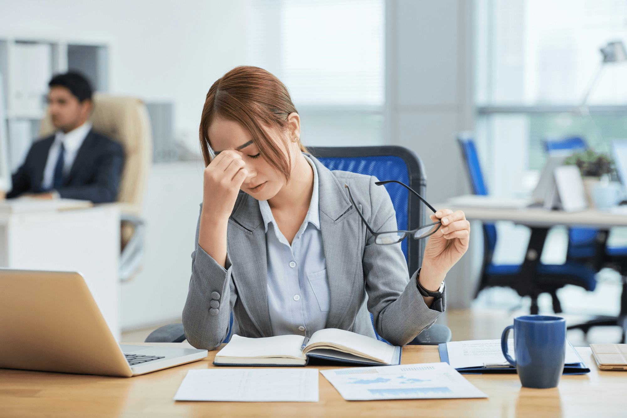 Working woman feeling stressed at the office desk, holding glasses, indicating how stress contributes to hormonal imbalance and unwanted weight gain