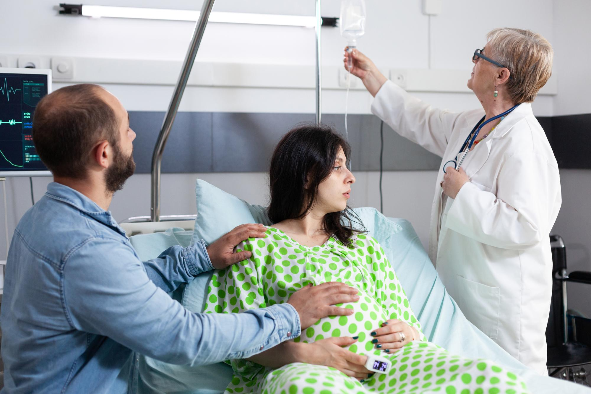 A pregnant woman lies in a hospital bed, hooked up to monitors, as her doctor and partner support her.