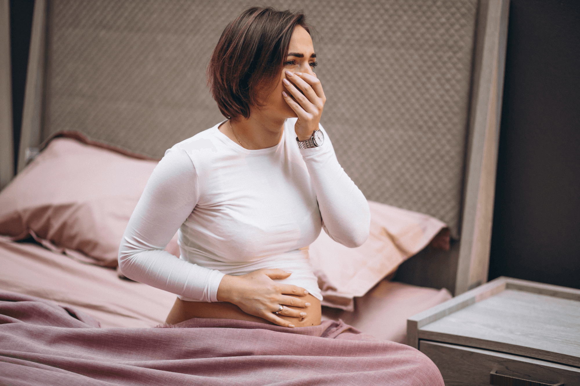 Pregnant woman sitting on bed feeling sick with symptoms of nausea during pregnancy from gestational diabetes and fatigue at home.