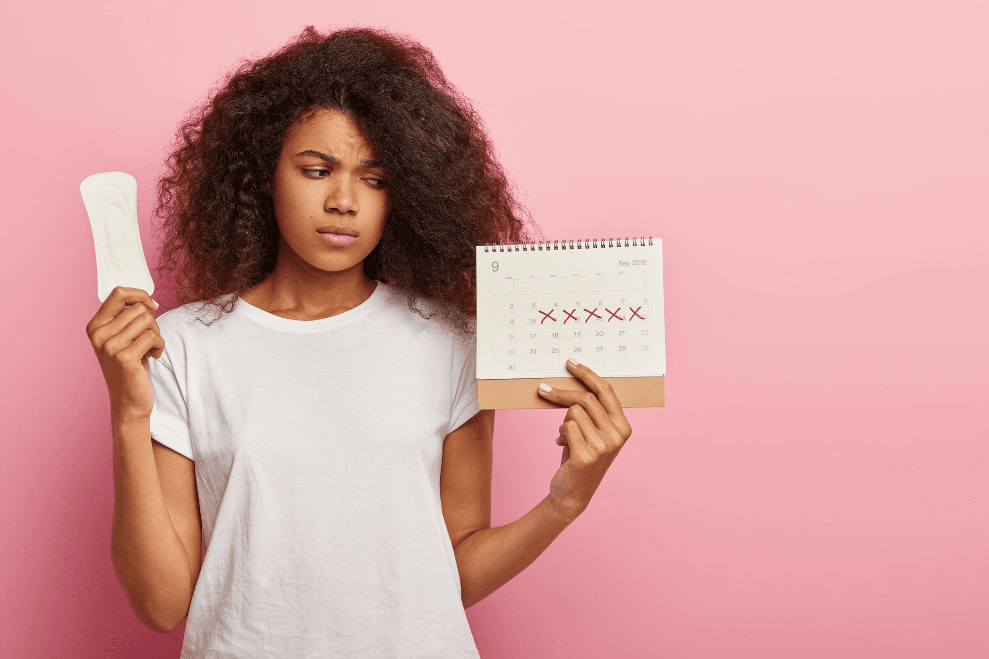A woman in her room holding a sanitary pad and a calendar while feeling confused and unhappy.