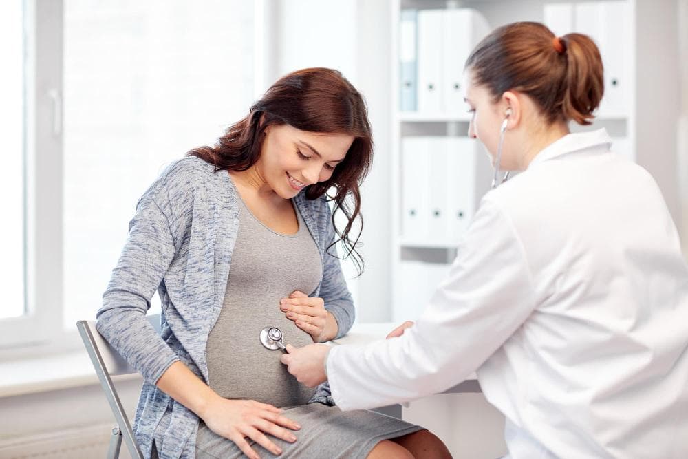 A joyful, pregnant woman is seated in a chair and has her hands placed gently upon her bump. A health professional wearing a white coat is using a stethoscope and listening to the baby's heartbeat.