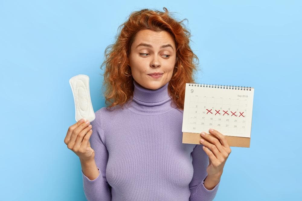 A young woman with curly red hair is joyfully playing with a sanitary pad in one hand, and a calendar showing marked dates of a menstrual period with red X marks in the other. The background is in soft blue colors.