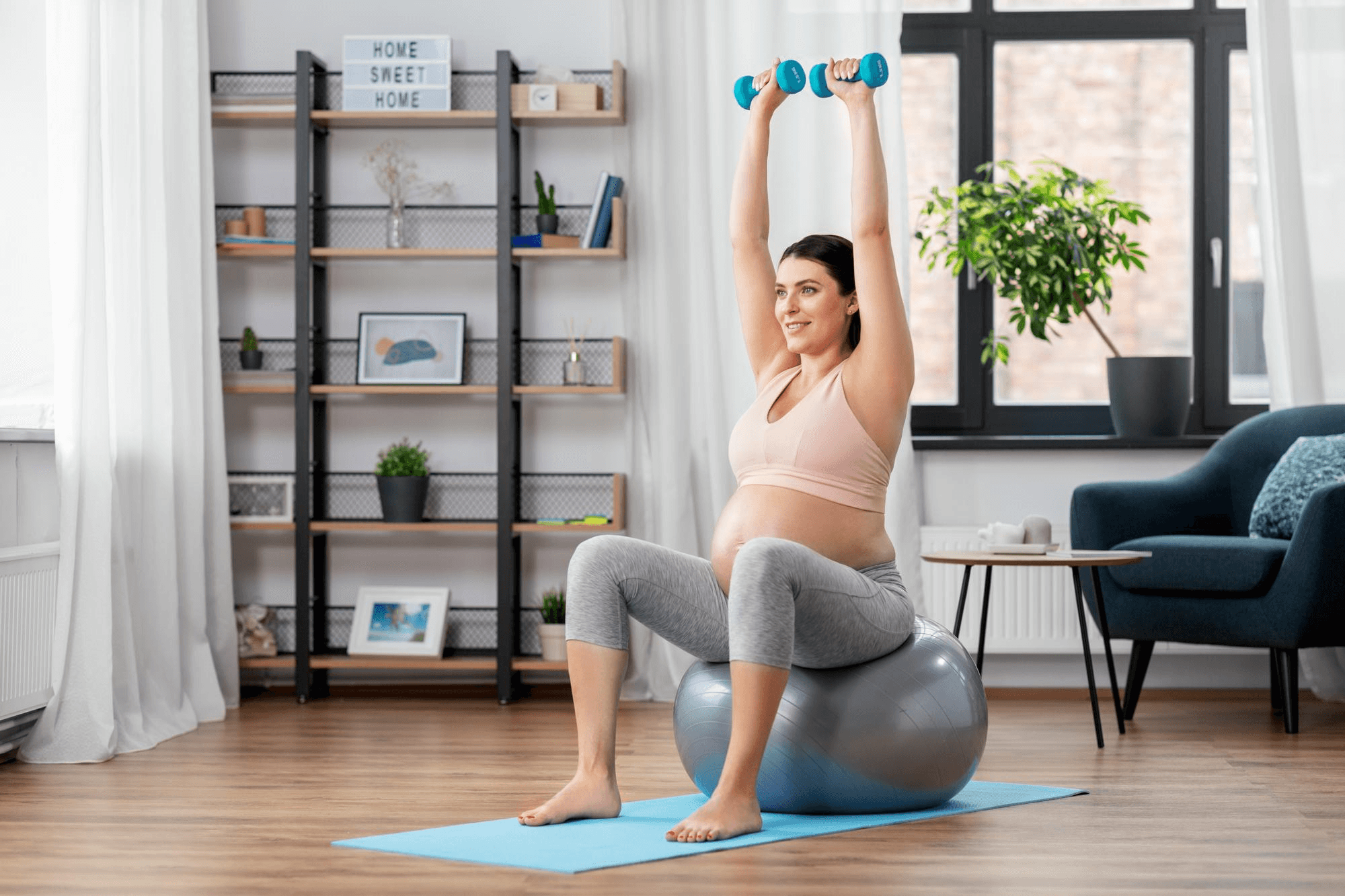 A pregnant woman is exercising by sitting on a yoga ball and is holding and lifting two dumbbells in each hand. She is smiling and the background is her bedroom.