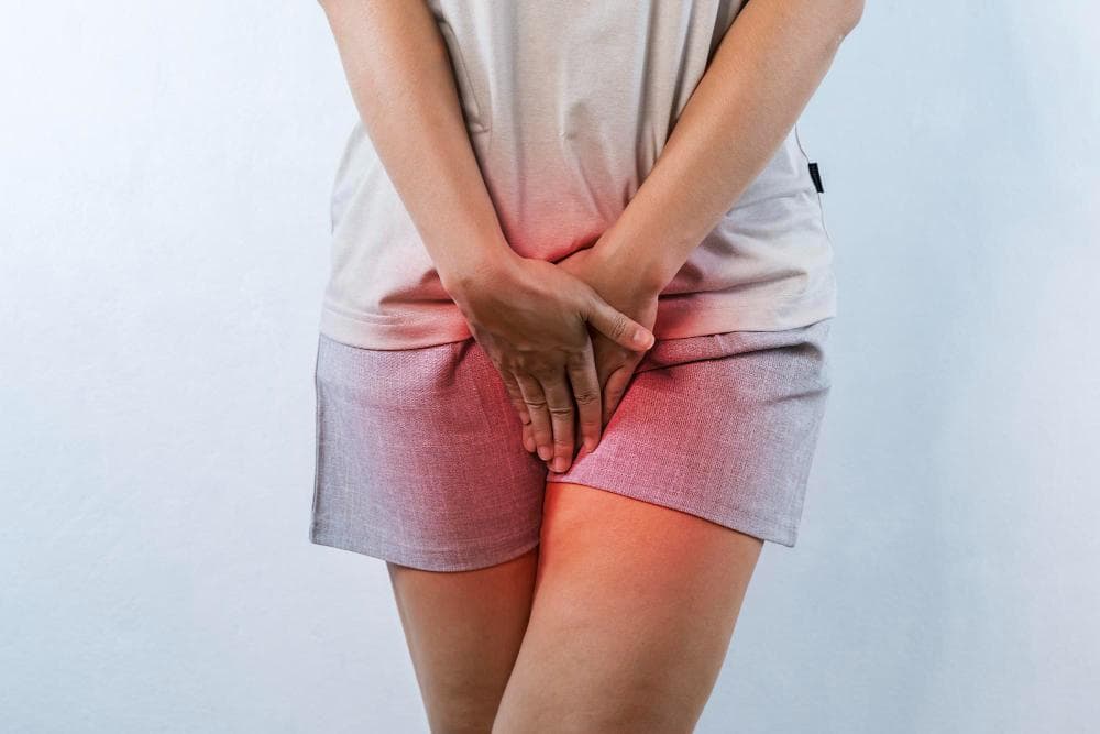 A woman is standing with her legs crossed sighting pain or irritation and her hands placed over her vagina, conveying discomfort.