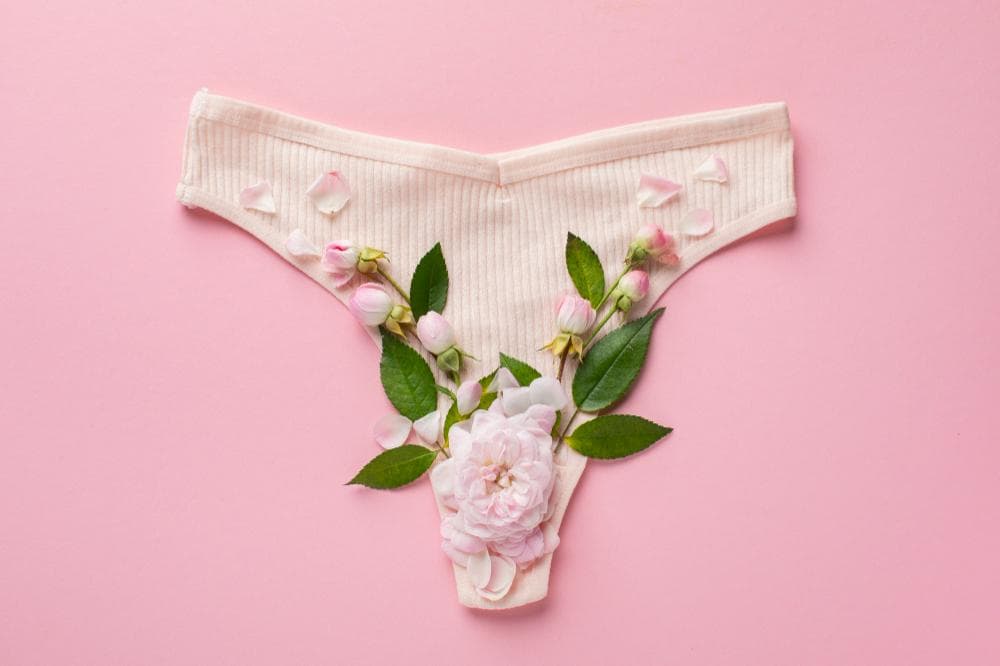A pair of women's panties is creatively decorated with a variety of pink and white flowers and green leaves. The panties are placed on a soft pink background.