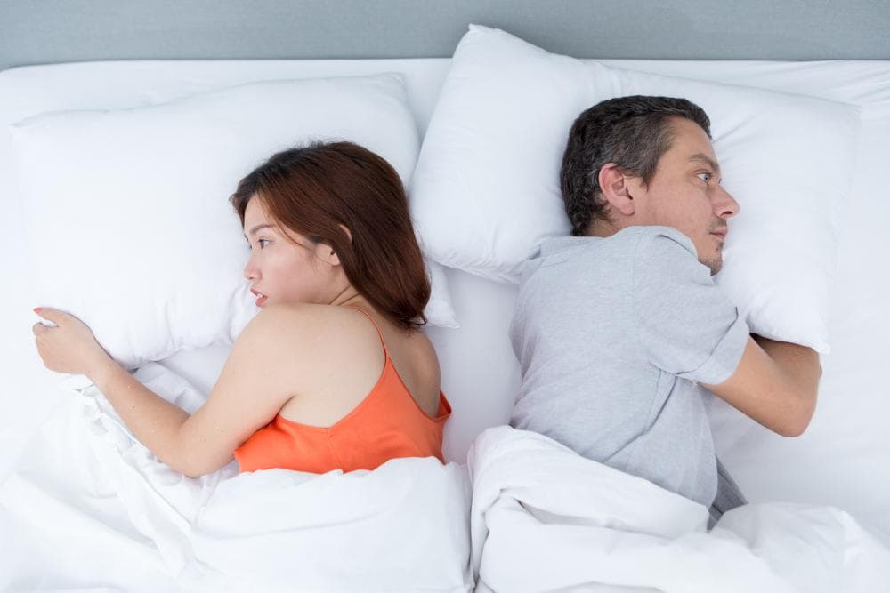 A couple is lying in bed, facing away from each other with expressions of distress. The woman, wearing an orange top, looks pensive while the man, in a gray shirt, appears frustrated. They are separated by pillows.