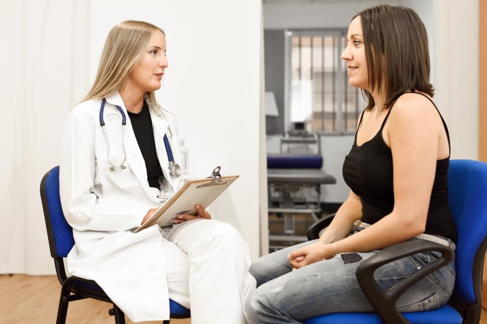 A female doctor, wearing a white coat and a stethoscope, is seated across from a female patient. The doctor is holding a clipboard and appears to be listening attentively. The environment is a clinical setting, suggesting a medical consultation.
